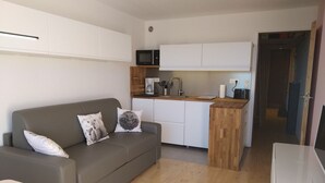 Un espacio habitable pequeño con un sofá gris, una mesa de centro de madera, una kitchenette con microondas y un mueble pequeño.