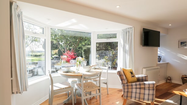 A bright dining area with a table set for two, surrounded by white chairs, a plaid sofa with a yellow pillow, and a view of greenery outside. - Old Kiln Cottage (Dumfries)