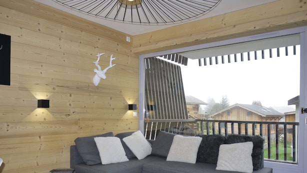 Ett modernt vardagsrum med en grå hörnsoffa, två vita, fluffiga pallar, ett vitt soffbord och ett stort fönster med utsikt över hus.
