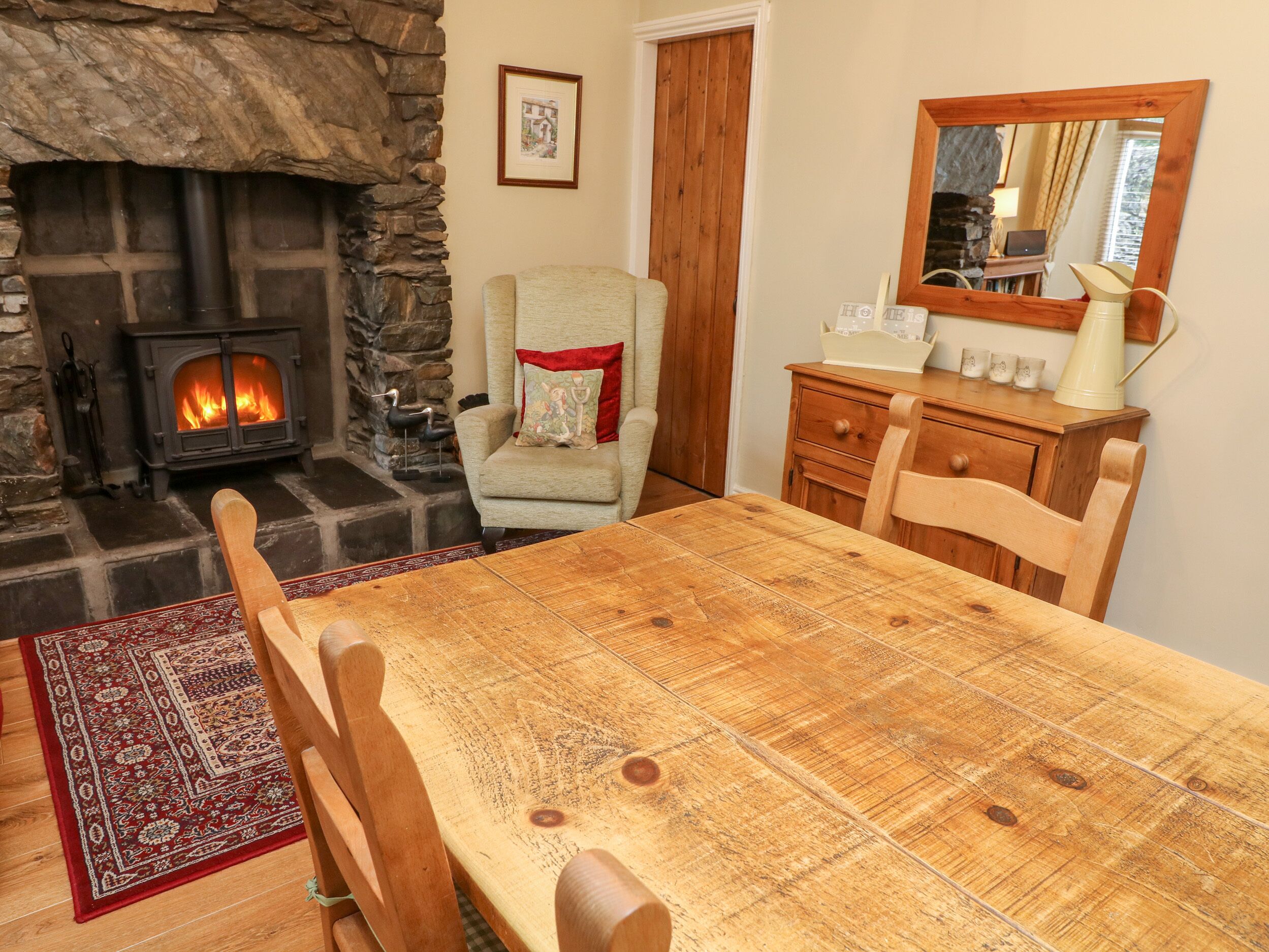 A cozy room with a stone fireplace, a dining table, wooden chairs, a small armchair, and a dresser with a mirror.