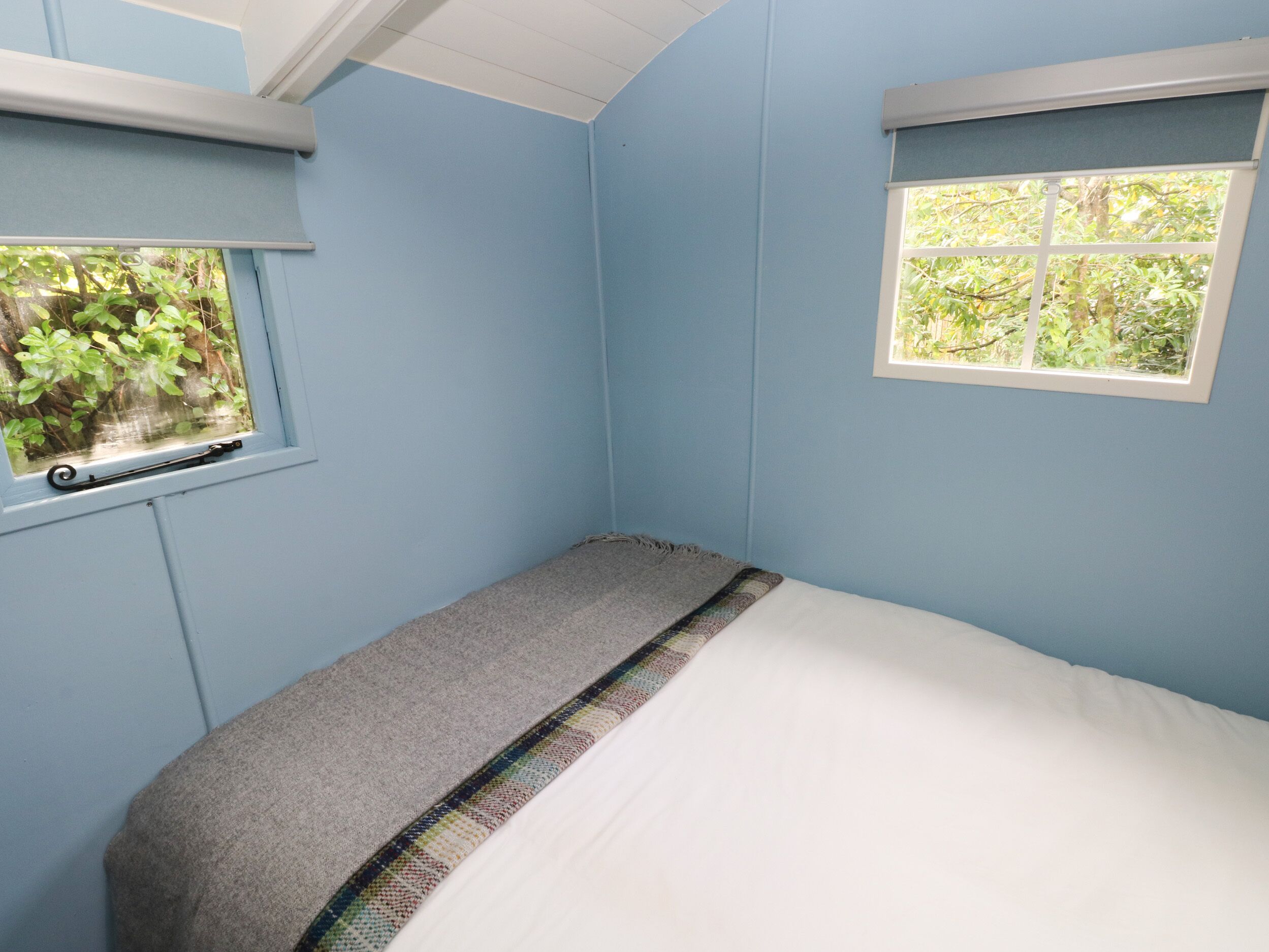 Une pièce aux murs bleu clair, avec un lit simple doté d’un matelas blanc et d’une couverture grise, une petite fenêtre donnant sur un paysage verdoyant et un plafond blanc.
