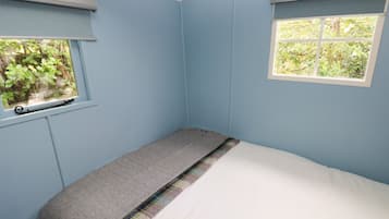 Une pièce aux murs bleu clair, avec un lit simple doté d’un matelas blanc et d’une couverture grise, une petite fenêtre donnant sur un paysage verdoyant et un plafond blanc.