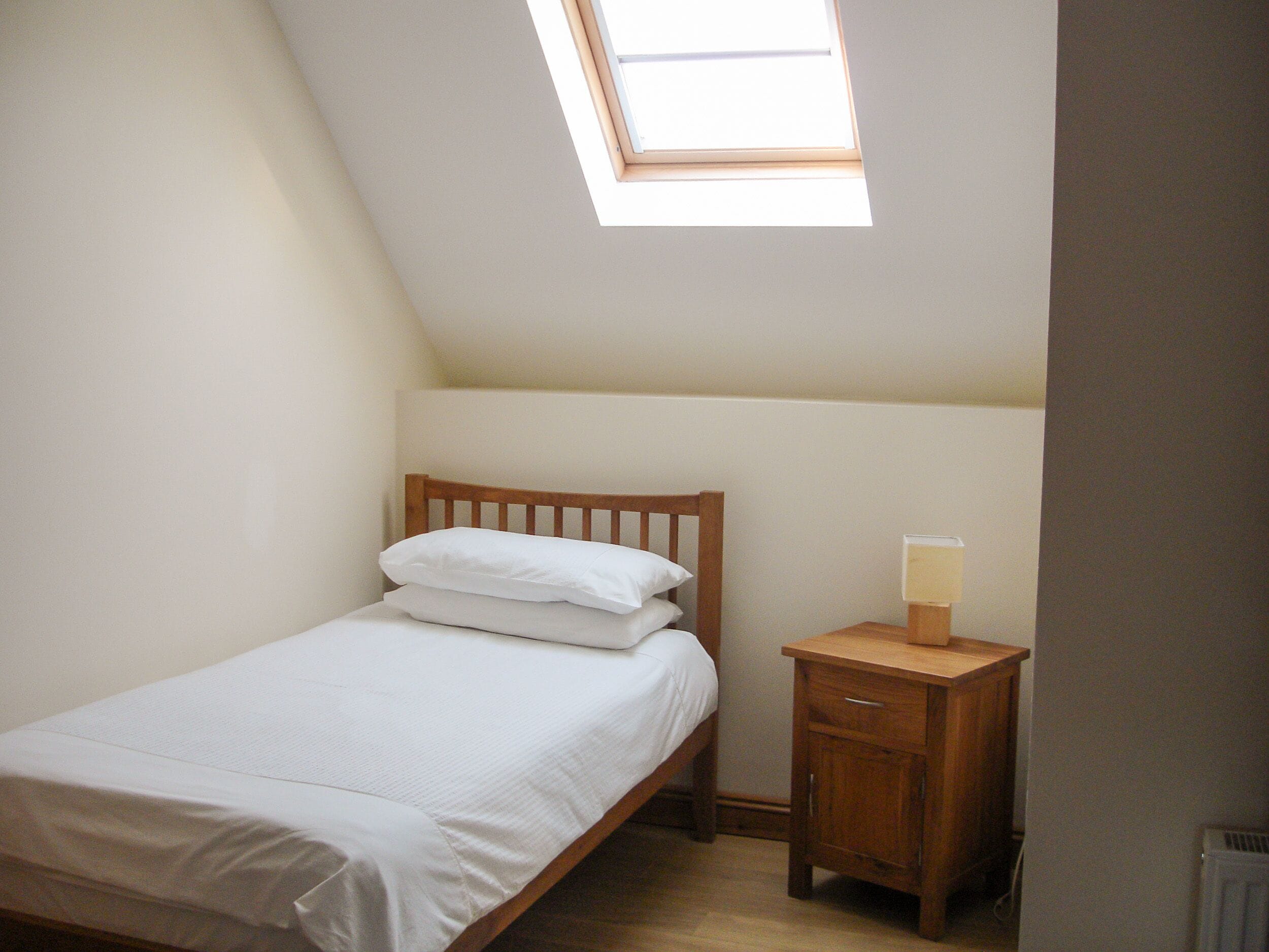 A small, neatly made bed with white linens in a room with a sloped ceiling and a skylight.