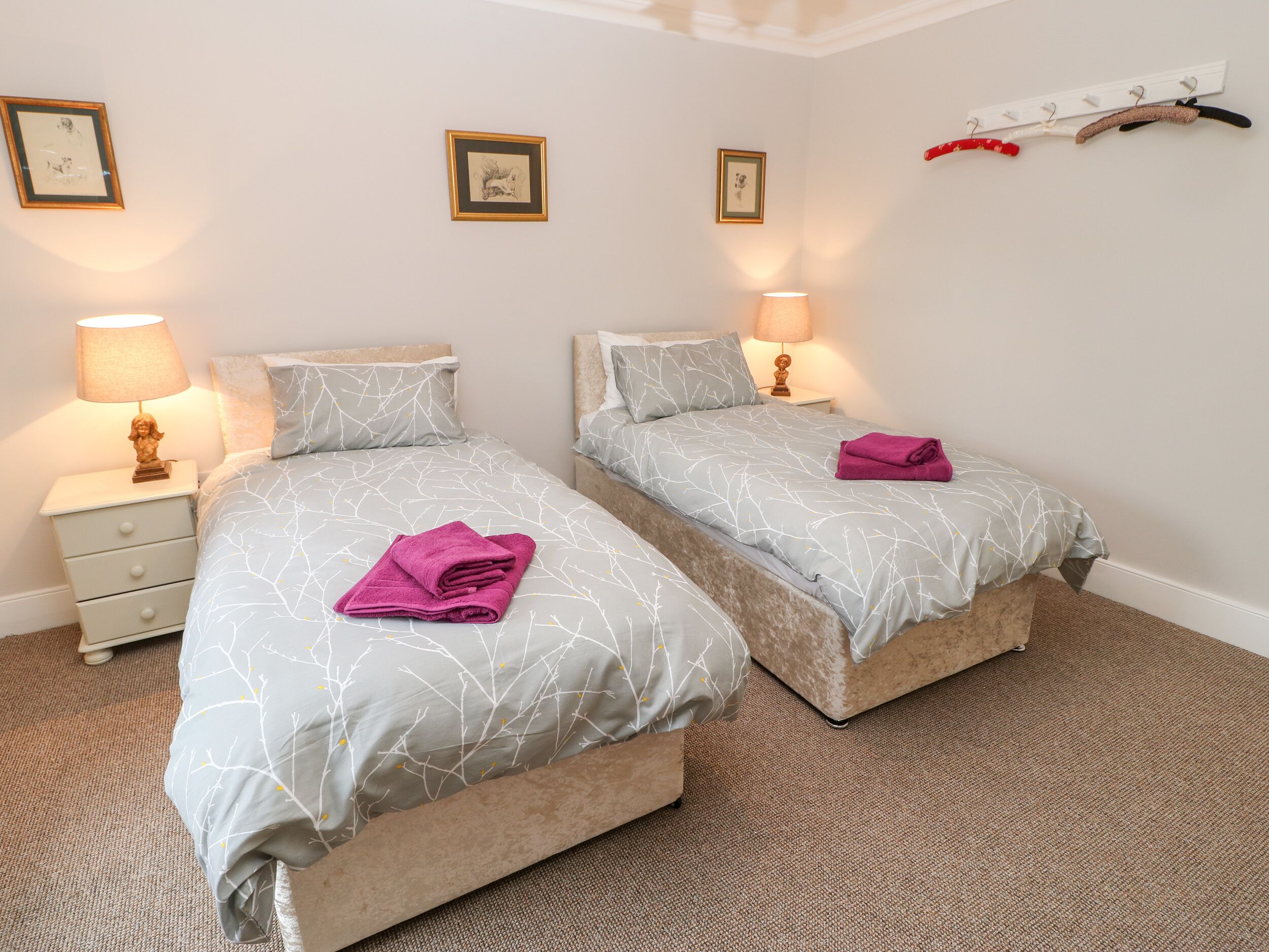 A double room with two beds, each with a grey patterned bedspread and pillows, a small bedside table with a lamp, and a wall-mounted coat rack.