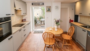 A modern kitchen with wooden cabinets, a built-in oven, and a dining area with a round table and chairs. - Hawthorne Cottage (Barnard Castle)