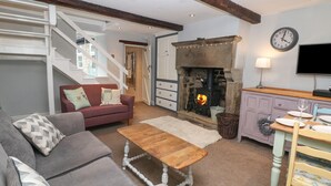 A cozy living room with a fireplace, a sofa, a coffee table, and a dining area with a table set for two. - Millstream Cottage (Hope Valley)