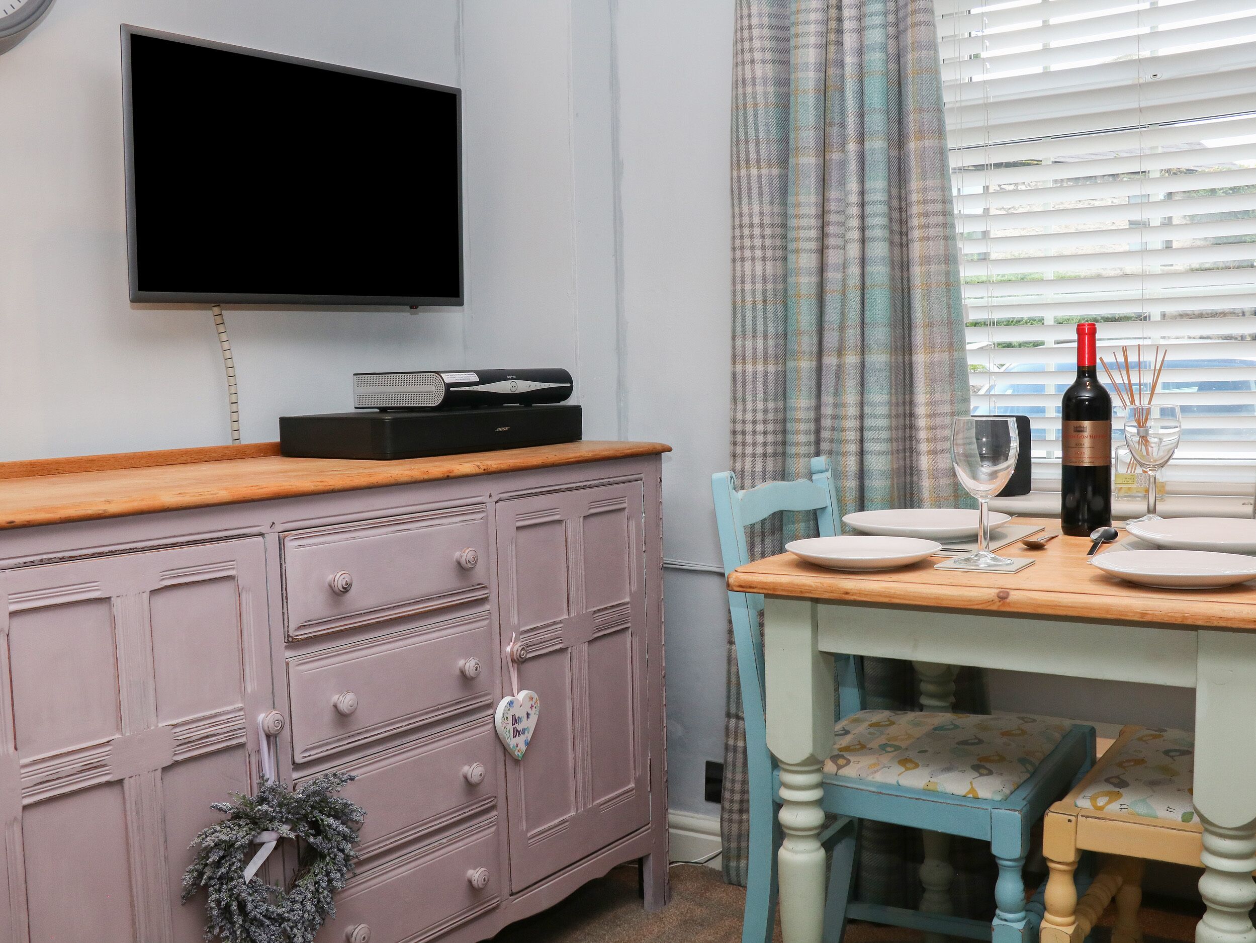 A dining area with a wooden table set for two, a television mounted on the wall, and a window with blinds.