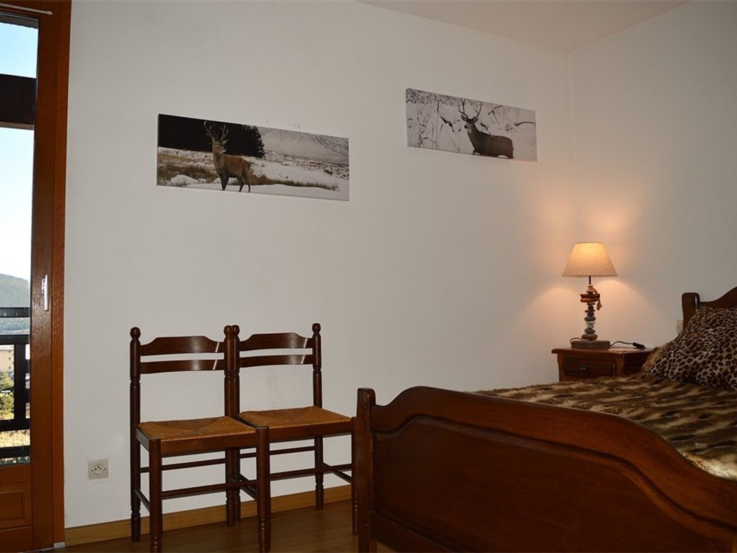 A bedroom with a wooden bed, a chair, a nightstand with a lamp, and two deer pictures on the wall.