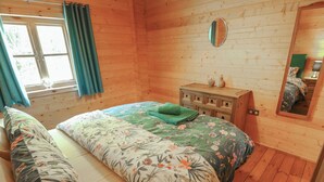 A wooden cabin room with a bed, a dresser, a mirror, and a window with curtains.