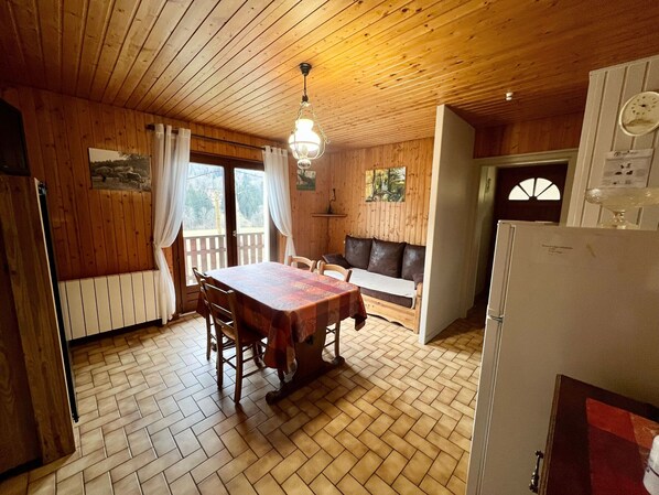 A wooden cabin interior with a dining table, chairs, a sofa, and a refrigerator.