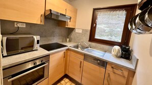 A compact kitchen with a microwave, stove, sink, and a window with a view of trees. - Chalet en Forêt Hautes-Vosges avec Terrasse, Proche Gérardmer, 2 Chambres, WiFi, Équipements Complets (Rochesson)