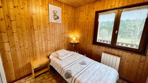 A wooden-paneled room with a bed, a bench, a wall-mounted lamp, and a window with a view of greenery. - Chalet en Forêt Hautes-Vosges avec Terrasse, Proche Gérardmer, 2 Chambres, WiFi, Équipements Complets (Rochesson)