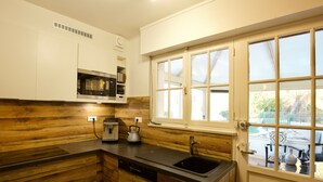 A compact kitchen with wooden cabinetry, a built-in microwave, a kettle, and a window with blinds. - Magnificent Retreat in Vosges: Sauna, Hot Tub, Park, and More! (Colroy-la-Grande)