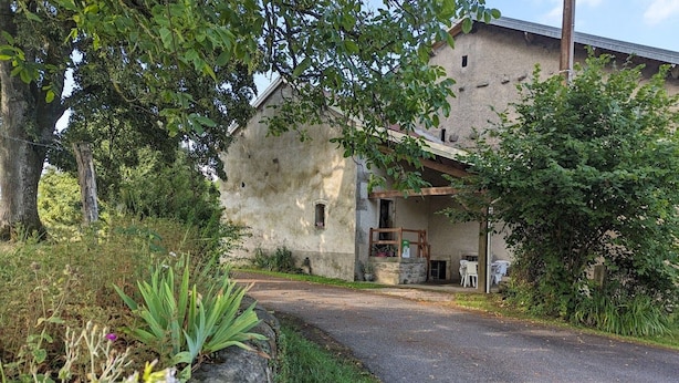 A rustic stone building with a wooden porch, surrounded by greenery and a paved driveway.