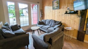 A cozy living room with a grey sofa set, a small round table, and a television mounted on a wooden wall. - Spacious Rustic Cottage with Terrace, Close to Gérardmer – Ideal for Families (Granges-Aumontzey-Aumontzey)