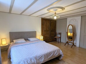 A bedroom with a large bed, a wooden wardrobe, a chair, a mirror, and a ceiling light fixture.