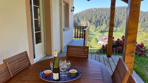 A wooden table with a bottle of wine, glasses, and small bowls on a patio with a scenic view of a forested area and buildings.