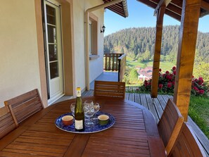 A wooden table with a bottle of wine, glasses, and small bowls on a patio with a scenic view of a forested area and buildings. - Spacious chalet with balcony, free WiFi, close to lake and ski resort (Gérardmer)