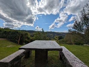 A stone picnic table in a grassy area with a view of a forested hill and a partly cloudy sky.