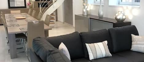 Una sala de estar moderna con un sofá negro, una zona de comedor con una mesa larga y una escalera de caracol.