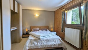 Ein Schlafzimmer mit hölzernem Kopfteil, einem Bett mit weißer Bettwäsche, zwei Nachttischlampen, einem Fenster mit Vorhängen und einem Heizkörper.