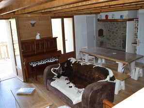A rustic cabin interior with a wooden dining table, a sofa with a cowhide cover, a piano, and a stone wall.