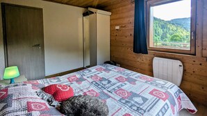 Quarto com teto de madeira, cama com colcha estampada, radiador e janela com vista para vegetação.