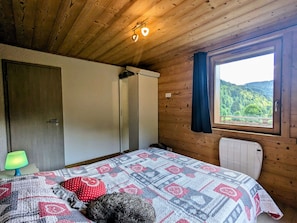 A bedroom with a wooden ceiling, a bed with a patterned quilt, a radiator, and a window with a view of greenery.