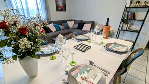 A dining area with a white table set for four, a vase of flowers, and a bookshelf in the background. - Cozy Retreat for 4 with South-Facing Balcony, Garage, and Easy Access to Nature and Local Attractions (Gérardmer)
