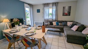 A living room with a dining area, a sofa set, a TV, and a painting on the wall. - Cozy Retreat for 4 with South-Facing Balcony, Garage, and Easy Access to Nature and Local Attractions (Gérardmer)
