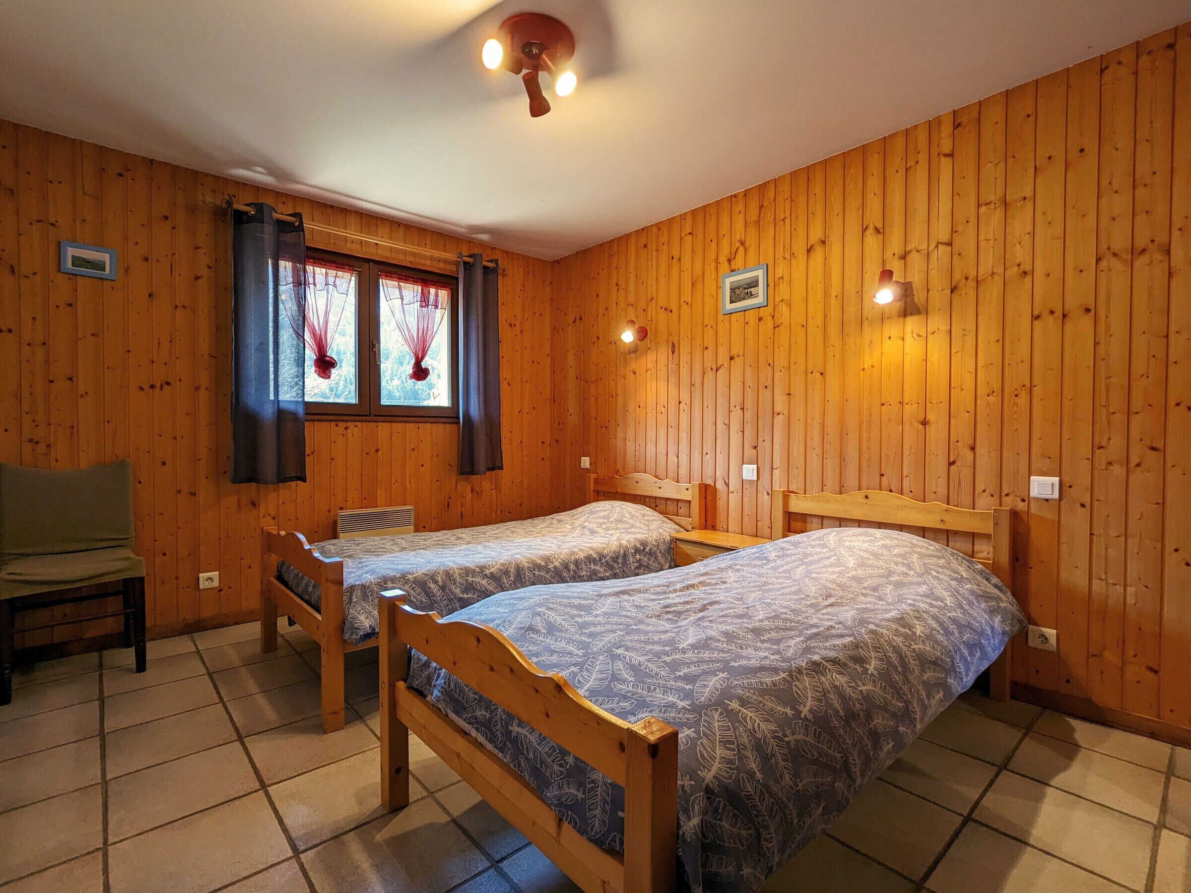 A room with wooden walls, two single beds with patterned bedding, a wooden chair, a table, and a window with curtains.
