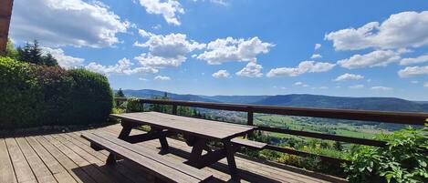 Une terrasse en bois avec une table de pique-nique et une rambarde donnant sur un paysage magnifique, avec des montagnes et un ciel dégagé.