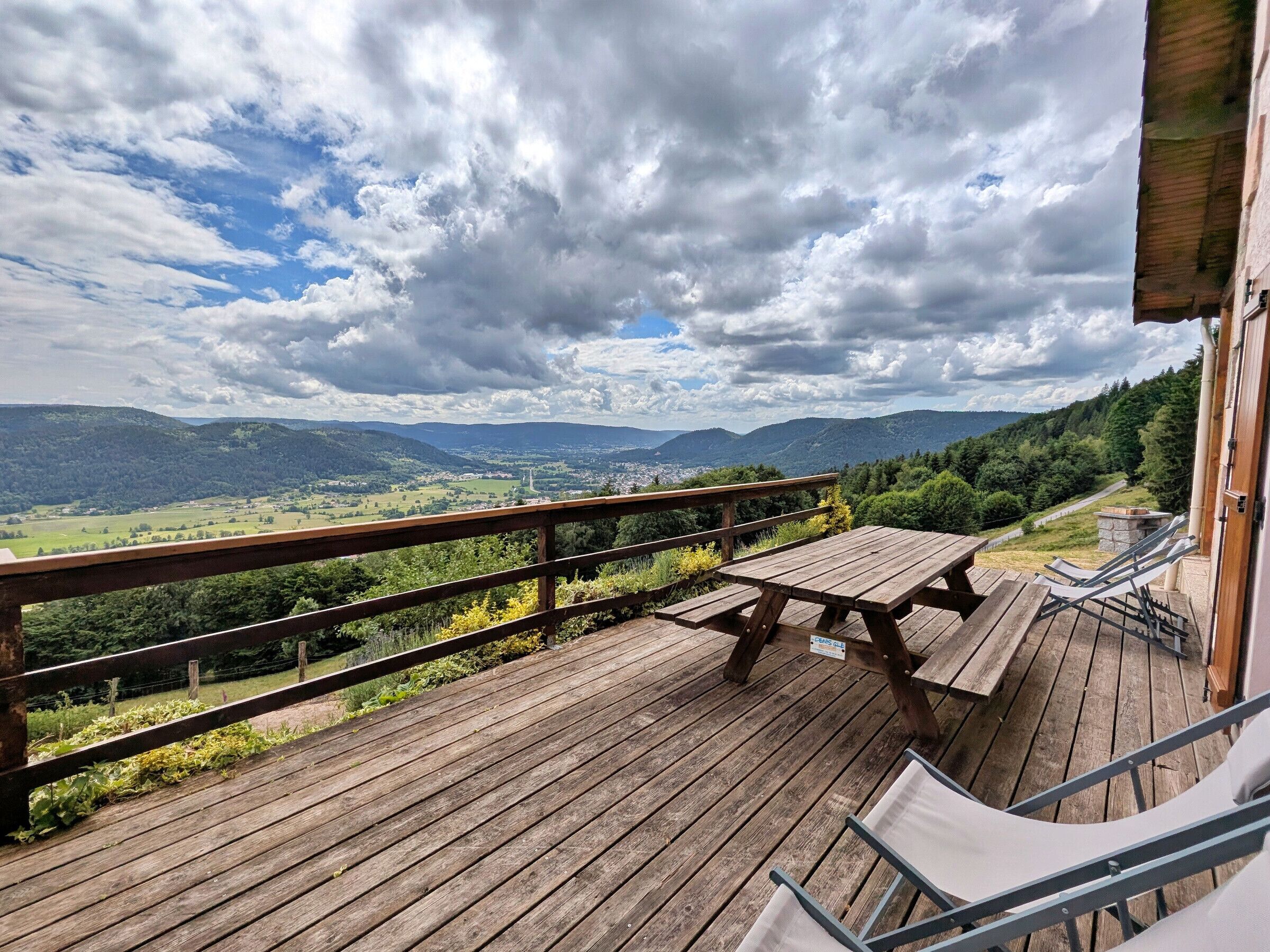 Eine Holzterrasse mit Picknicktisch und Stühlen, mit Blick auf ein Tal mit Bergen und einer Stadt.
