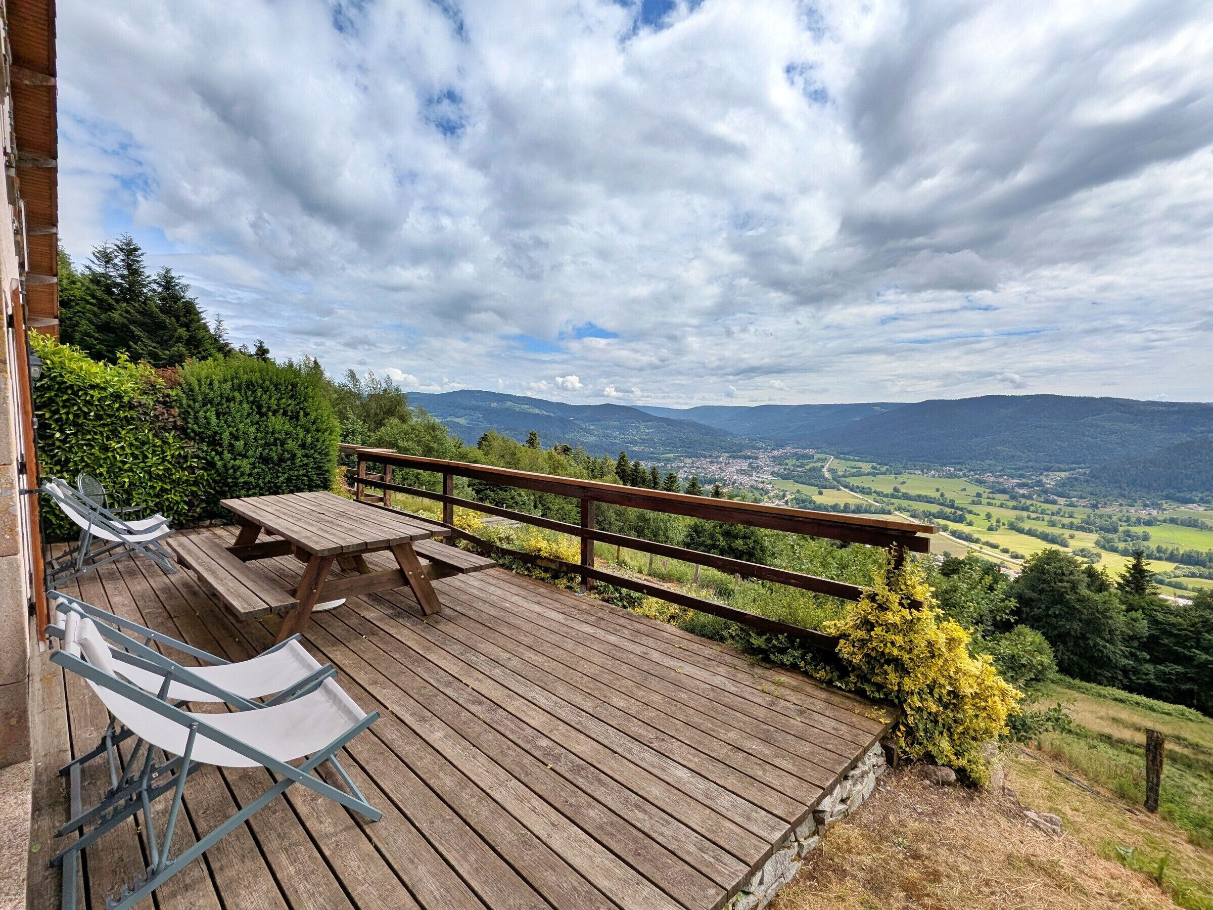 A wooden deck with a table and chairs, overlooking a valley with mountains and a town.