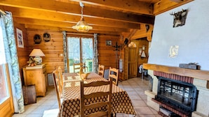 A cozy wooden cabin with a dining table set for four, a fireplace, and a large window offering a view of the outdoors. - Cozy Chalet Near Ski Slopes and Lake, Sleeps 7, WiFi, Fireplace, Large Garden, Garage (Le Menil)