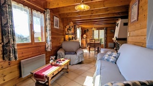 A cozy cabin interior with a wooden floor, a sofa, a chair, a table with a red cloth, and a hanging light fixture. - Cozy Chalet Near Ski Slopes and Lake, Sleeps 7, WiFi, Fireplace, Large Garden, Garage (Le Menil)
