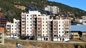 Un edificio residencial de varios pisos con balcones, ubicado en una zona montañosa con un bosque denso.