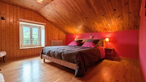 Ein Schlafzimmer mit geneigter Decke, Holzwänden, einem Bett mit rosa und braunen Textilien, einem Nachttisch mit Lampe und einem Fenster mit Blick auf Grünflächen.