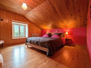 Ein Schlafzimmer mit geneigter Decke, Holzwänden, einem Bett mit rosa und braunen Textilien, einem Nachttisch mit Lampe und einem Fenster mit Blick auf Grünflächen.
