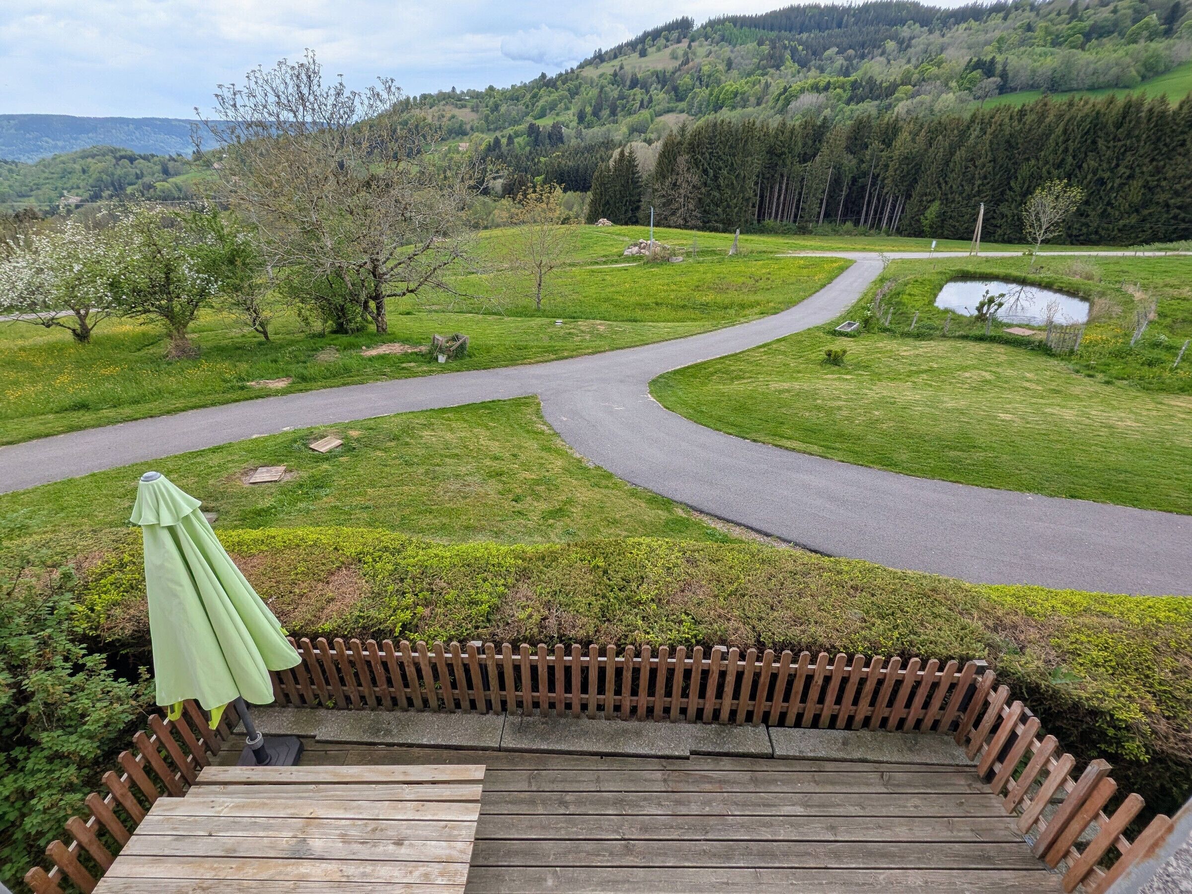 Eine Holzterrasse mit einem grünen Sonnenschirm, mit Blick auf eine kurvenreiche Straße und eine grüne Landschaft mit Bäumen und einer Hügelkette in der Ferne.