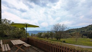Eine Holzterrasse mit Tisch und Sonnenschirm, umgeben von einem Holzzaun, mit Blick auf eine grüne Landschaft aus Bäumen und Hügeln.