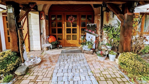 A rustic building entrance with a wooden porch, a stone pathway, and potted plants. - Charming Renovated Farmhouse, Near Ski Resorts & Hiking Trails, All-Inclusive, Close to Amenities (Le Syndicat)