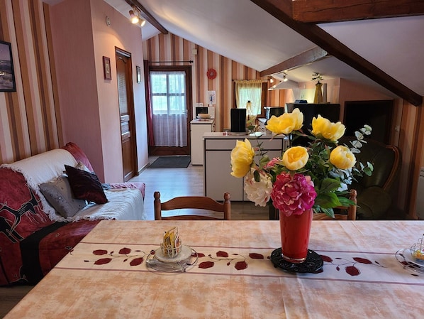 A cozy living room with a sofa, a dining table with a vase of flowers, and a kitchen in the background.