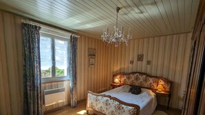 Une chambre avec un plafond en bois, un lustre, un lit recouvert d’un tissu à motifs floraux, une table de chevet avec une lampe et une fenêtre avec des rideaux.