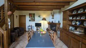 Un espace repas comprenant une longue table, des chaises, une cheminée et des étagères en bois où sont rangés de la vaisselle.
