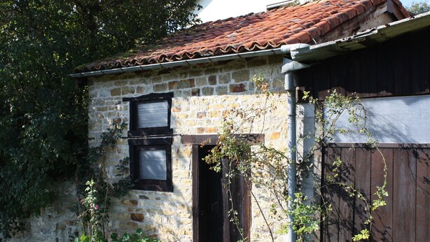 Un bâtiment en pierre avec un toit en tuiles, une porte en bois et un petit jardin agrémenté de plantes à fleurs.