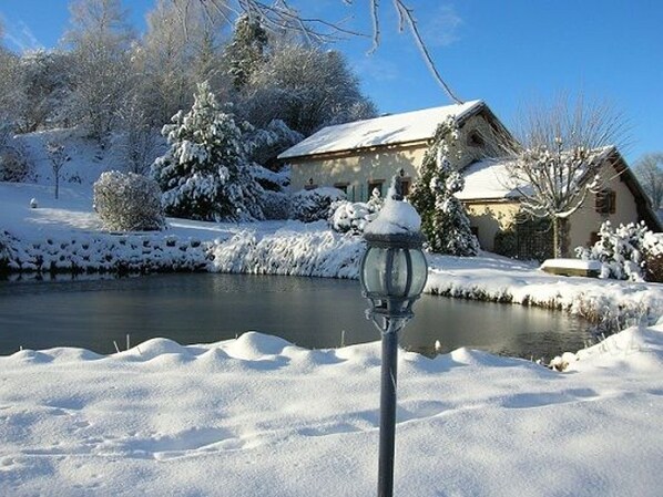 A snow-covered landscape with a house and a pond.