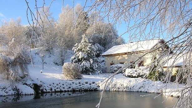 Un paysage hivernal paisible, avec une nature enneigée, un petit étang et une maison au toit en pente.