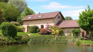 Une maison en pierre avec un toit de tuiles, un étang et une végétation luxuriante.