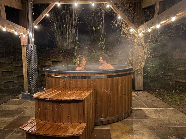 A hot tub with a wooden exterior, surrounded by stone flooring and wooden beams.